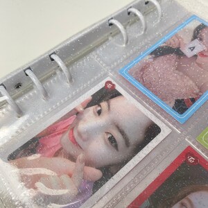 May include: A clear plastic binder with silver glitter embedded in the plastic. The binder has a ring mechanism and is filled with photo cards. The photo cards feature a young woman with long dark hair and a pink shirt. The photo cards have a red heart with the word "twice" written on them.