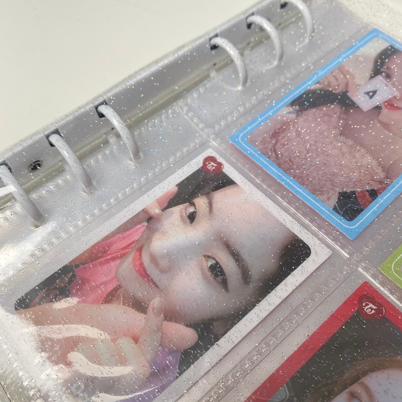 May include: A clear plastic binder with silver glitter embedded in the plastic. The binder has a ring mechanism and is filled with photo cards. The photo cards feature a young woman with long dark hair and a pink shirt. The photo cards have a red heart with the word "twice" written on them.