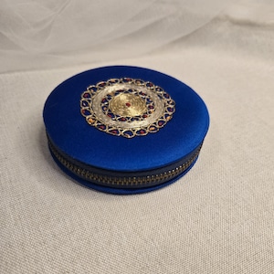 May include: A round, cobalt blue jewelry box with a gold and silver filigree design on the lid. The filigree is embellished with red and amber-colored stones. The box has a brass zipper.
