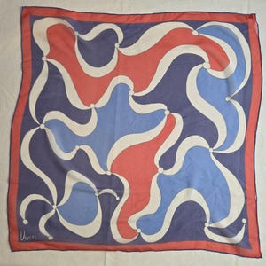 May include: Square silk scarf with a geometric design in red, blue, and white. The scarf has a navy blue background with a red border. The design features curved lines and circular accents. The scarf has the signature "Ugnia" in the bottom left corner.