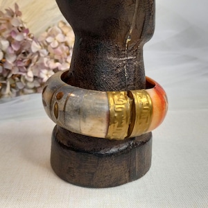 May include: A silver and gold bangle bracelet with a red and white design. The bracelet is on a wooden display stand.