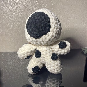 May include: A handmade crochet doll in a standing position. The doll is primarily white with black spots and a large black circle on its head. The doll has small arms and legs, and is made of a soft, textured yarn.