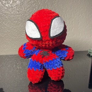 May include: A red and blue crocheted plush toy shaped like Spiderman. The toy has a black spider symbol on its chest and white eyes.