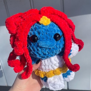 May include: A handmade crochet doll featuring a blue face, red hair, and a white and yellow outfit. The doll has large black eyes and a small yellow accent on its forehead. The plushie is made of soft, textured yarn.