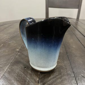 May include: A ceramic pitcher with a gradient design, transitioning from black at the top to light blue at the base. The pitcher has a curved handle and a spout for pouring. The handmade pottery is on a wooden surface.