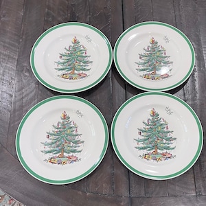 May include: Four white ceramic plates with a green border. Each plate features a detailed illustration of a decorated Christmas tree with colorful ornaments and presents at the base. The plates are arranged on a dark wooden surface.