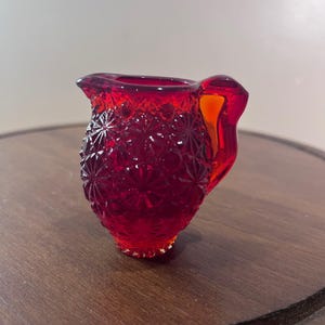 May include: A small, vibrant red glass creamer pitcher featuring a raised, geometric starburst pattern across its exterior. The pitcher has a curved handle on the right side and a distinct pouring spout, showcasing a translucent, deep ruby red color.