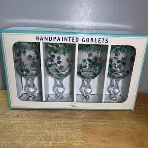 May include: A set of four clear glass goblets in a rectangular box labeled HANDPAINTED GOBLETS. Each goblet features a hand-painted design of green leaves and small purple flowers around the rim and bowl, supported by a clear glass stem and base.
