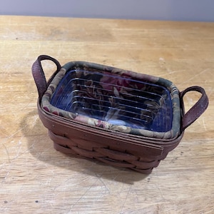 May include: A brown woven basket with two leather handles. The basket has a floral fabric lining and a clear plastic insert. The basket is rectangular and sits on a wooden surface.