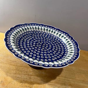 May include: An oval ceramic serving platter with a scalloped edge, featuring a dense pattern of small blue and brown circular motifs on a white background, accented by a border of green and blue leaf-like shapes around the inner rim.