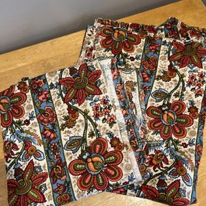 May include: A set of floral patterned fabric, featuring large red and orange flowers with green and blue accents on an off-white background. The fabric is arranged in a folded stack and a pair of pants.