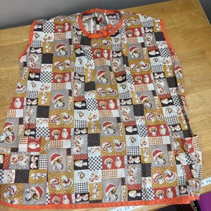May include: A colorful apron with a patchwork design featuring mushroom illustrations. The apron has an orange trim and neck tie. The fabric incorporates squares of various patterns and colors, including brown, tan, and black.