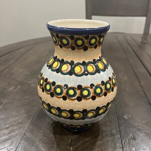 May include: A small, white ceramic vase with a blue rim and a pattern of yellow and black circles. The vase is sitting on a wooden table.