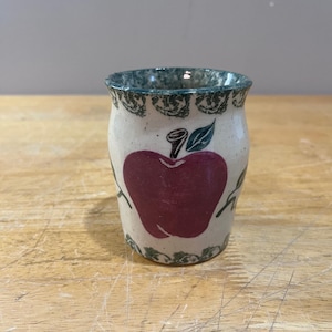 May include: A small, ceramic jar with a cream-colored base and a dark red apple design. The rim and base feature a green, speckled pattern. Green leaves and stems accent the apple. The jar is likely used for kitchen storage or decoration.