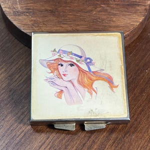 May include: A square metal compact mirror featuring a printed illustration of a person with long orange hair wearing a white sun hat adorned with a blue ribbon and small pink flowers. The compact has a silver-toned frame and two metal latches.