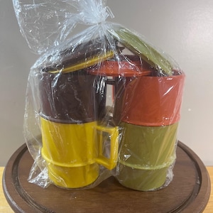 May include: A set of four stackable plastic mugs in brown, yellow, orange, and olive green, each featuring a side handle and a matching lid. The mugs are bundled together inside a clear plastic bag, showing a cylindrical shape and smooth finish.