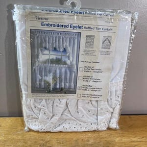 May include: A white embroidered eyelet ruffled tier curtain set in clear plastic packaging. The label displays the text Vienna Embroidered Eyelet Ruffled Tier Curtain and lists contents including a 60 inch by 15 inch double crescent valance.