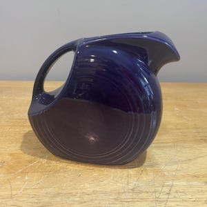 May include: A cobalt blue ceramic pitcher with a curved handle and a spout. The pitcher has a rounded body with concentric ridges and sits on a wooden surface. The pitcher is approximately 8 inches tall.