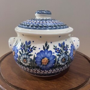 May include: A round ceramic lidded jar with two side handles, featuring a cream-colored base decorated with hand-painted blue flowers, green leaves, and small blue dots. The lid and base are bordered by a repeating pattern of blue and brown circles.
