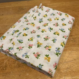 May include: A stack of white fabric napkins with a floral pattern. The pattern features small flowers in shades of pink, yellow, and blue, with green leaves. The napkins are neatly folded and stacked, ready for use.