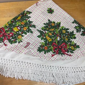 May include: A white circular tree skirt featuring a red and green grid pattern, decorated with printed holly, red bows, drums, bells, and nutcrackers. The edge is finished with a wide white crocheted trim and long fringe, measuring 33 inches.