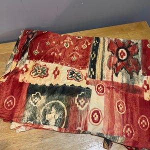 May include: A folded textile with a patchwork design. The fabric features a mix of red, cream, black, and green hues. The pattern includes floral and geometric shapes, with a vintage aesthetic. The textile is likely a scarf or a decorative piece.