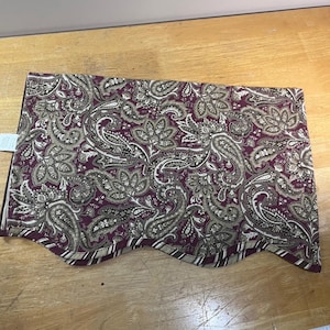 May include: A rectangular fabric valance featuring a deep burgundy background with a repeating paisley and floral pattern in shades of tan, beige, and dark brown. The bottom edge is scalloped and finished with a striped burgundy and white trim.