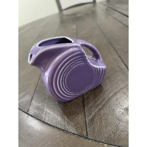 May include: A lavender-colored ceramic pitcher with a rounded body and a handle. The pitcher has a spout and a series of concentric rings that encircle the body. The pitcher is sitting on a wooden surface.