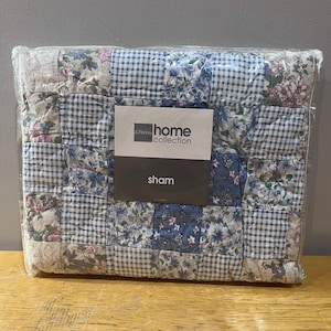 May include: A packaged sham from the JCPenney Home Collection. The sham features a patchwork design with squares of floral, gingham, and solid blue fabrics. The word "sham" is printed on the label.