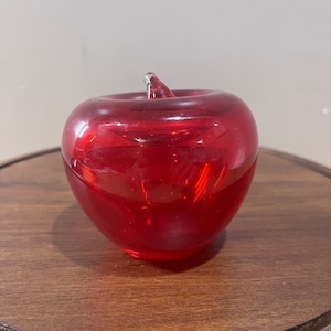 May include: A solid, translucent red glass apple figurine with a smooth, rounded body and a small, clear glass stem protruding from the top center. The object features a polished, reflective surface that catches light, creating deep red tones.