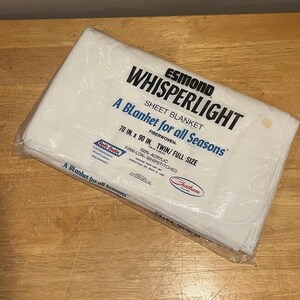 May include: A white Esmond Whisperlight sheet blanket, packaged in clear plastic. The text on the package reads "A Blanket for all Seasons" and "70 in. x 90 in. Twin/Full Size". The blanket is made of 100% acrylic fiber.