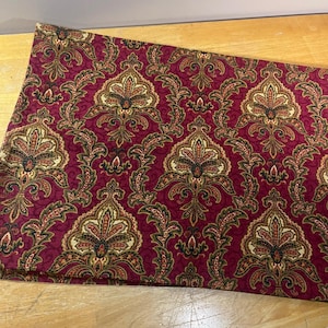 May include: A folded piece of fabric featuring a deep burgundy background with a repeating damask pattern in shades of gold, cream, and black. The intricate design consists of symmetrical floral and leaf motifs arranged in a vertical layout.
