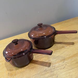 May include: Two vintage, matching, purple-tinted glass saucepans with lids. Each saucepan has a long, rectangular handle and a round lid with a small knob. The saucepans are sitting on a light-colored wooden surface.