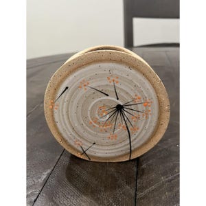 May include: A handmade ceramic vase with a beige exterior and a cream-colored interior. The interior features a spiral design with black lines and orange dots, resembling a dandelion. The vase is a unique piece of home decor.