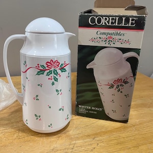 May include: White Corelle thermal server with a lid and handle, decorated with a red ribbon and holly berry design. The box next to it reads "Corelle Compatibles" and "Winter Holly".
