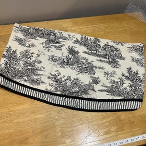 May include: A decorative valance featuring a black and white toile pattern with a striped border. The toile design depicts pastoral scenes with figures, trees, and buildings. The valance is made of fabric and is curved.