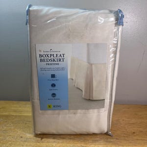 May include: A king-sized off-white boxpleat bedskirt packaged in a clear plastic bag. The product label features the text Home Classics, Boxpleat Bedskirt, Pristine, 15 in. Deep Skirt, Split Corners, Machine Washable, and K King.