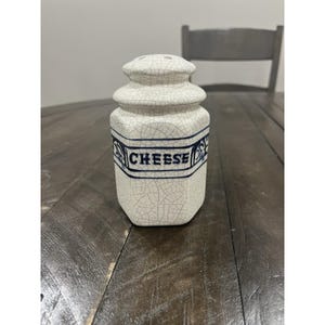 May include: A white ceramic cheese shaker with a crackle glaze finish. The shaker has a cylindrical shape with a rounded top and a blue band that reads "CHEESE". The shaker is sitting on a wooden table.