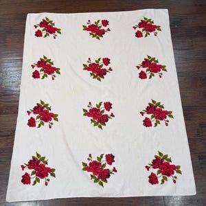 May include: White rectangular textile featuring a repeating pattern of red roses with green leaves. The roses are clustered in groups of flowers and leaves. The textile is likely a tablecloth or a decorative fabric.