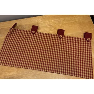 May include: A rectangular valance with a red and tan gingham pattern. The valance has several red fabric loops with button closures along the top edge. The valance is made of a soft fabric and is likely for a kitchen or dining room.