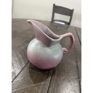 May include: A ceramic pitcher with a rounded body and a curved spout. The pitcher is a gradient of pink and green, with a handle on the side. The pitcher is likely vintage and suitable for serving beverages.
