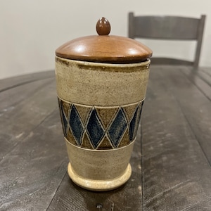 May include: A ceramic jar with a wooden lid. The jar has a beige background with a brown and blue diamond pattern.