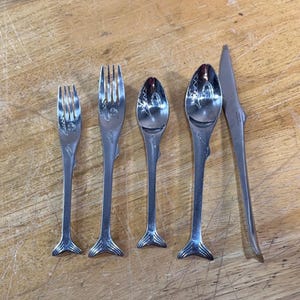 May include: A set of five stainless steel utensils featuring fish-shaped handles with engraved details. The collection includes two forks, two spoons, and one knife, all designed with a distinct tail-shaped base and etched patterns on the stems.