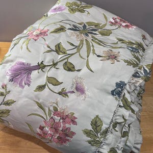 May include: A light blue floral comforter with embroidered flowers in shades of pink, purple, and green. The comforter has a ruffled edge and a decorative rope trim. The floral pattern is spread across the comforter.