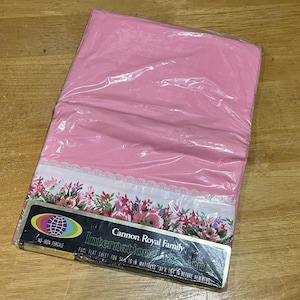 May include: A pink flat sheet in plastic packaging. The sheet has a floral border with pink, purple, and white flowers. The packaging includes the text "Cannon Royal Family International".