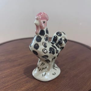 May include: A small ceramic figurine of a rooster with a glossy off-white finish, decorated with black polka dots across its body and tail. The rooster features a distinct pink comb and wattle, with small green markings at the base of the stand.