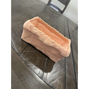 May include: A rectangular, light pink ceramic planter with a paper bag design. The interior is a slightly darker shade of pink. The planter is sitting on a dark wood surface.