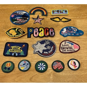 May include: A collection of fifteen embroidered fabric patches featuring various designs including a rainbow, a star with an American flag pattern, a peace sign, a snowflake, and text such as Camp Survivor, Pumpkin Patch, and I Love Brownie Girl Scouts.