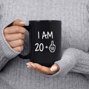 May include: Black coffee mug with white text that reads "I AM 20 +" with a middle finger emoji.