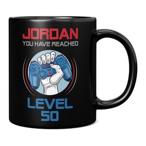 Könnte beinhalten: Schwarze Kaffeetasse mit einem weißen Design. Das Design zeigt einen blauen Videospielcontroller, der von einer Hand gehalten wird. Der Text "JORDAN YOU HAVE REACHED LEVEL 50" ist in Rot und Weiß.